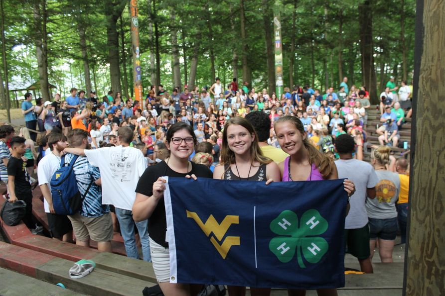 Three girls hold a flag with a WVU logo and 4-H logo in front of a large crowd of 4-Hers.